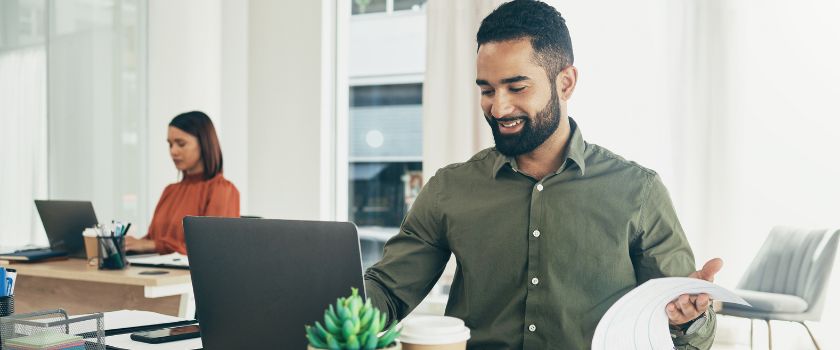 A smiling professional reviewing financial documents at his desk in a modern office. He is engaged with his work, with a laptop open in front of him and a relaxed, confident demeanor. In the background, a colleague is also working, creating a productive and collaborative atmosphere.